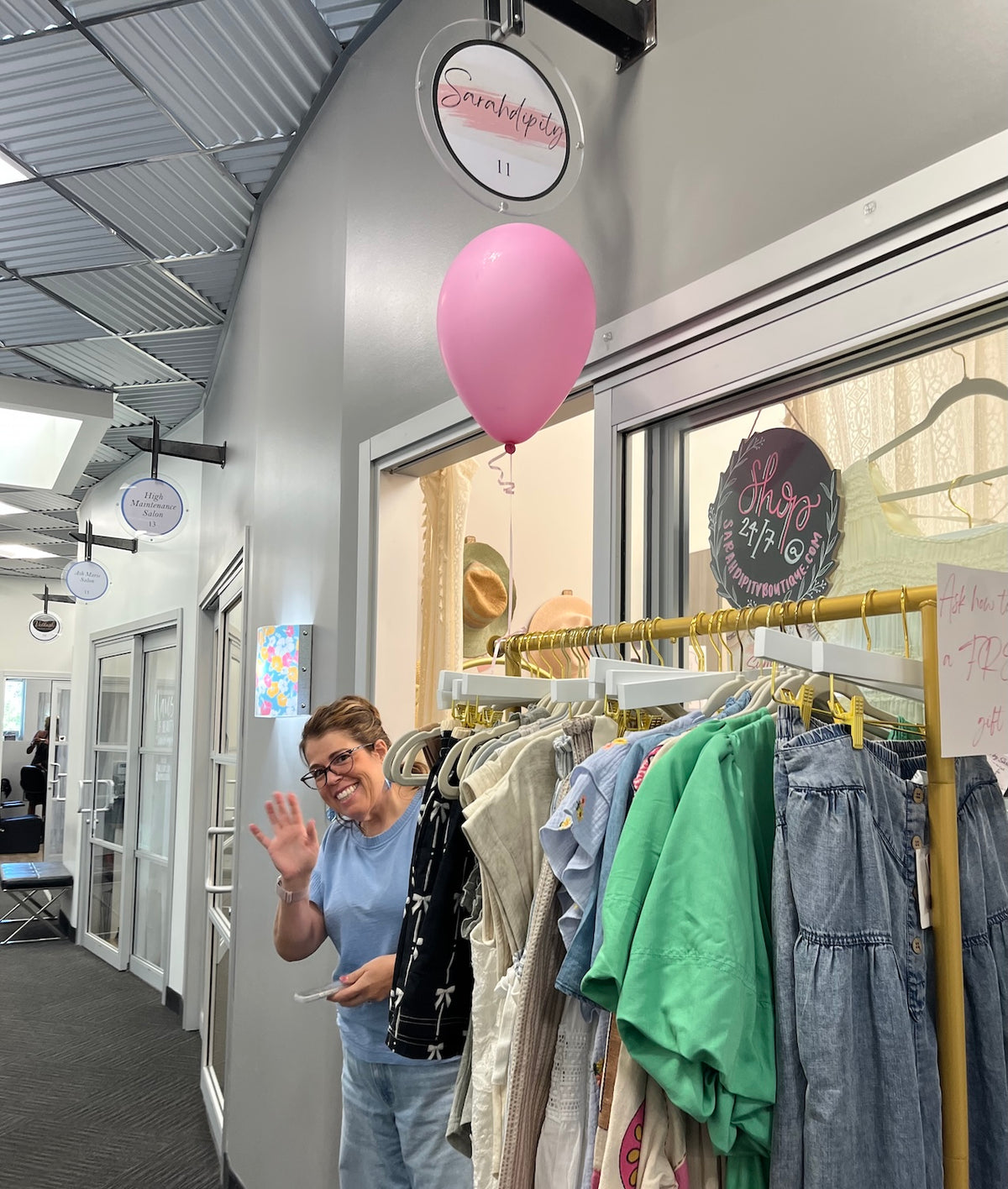 A woman waves from inside a boutique while standing next to a clothing rack filled with colorful garments, including a prominent green shirt and denim pieces. A pink balloon floats near the window, and shop signage is visible in the background.